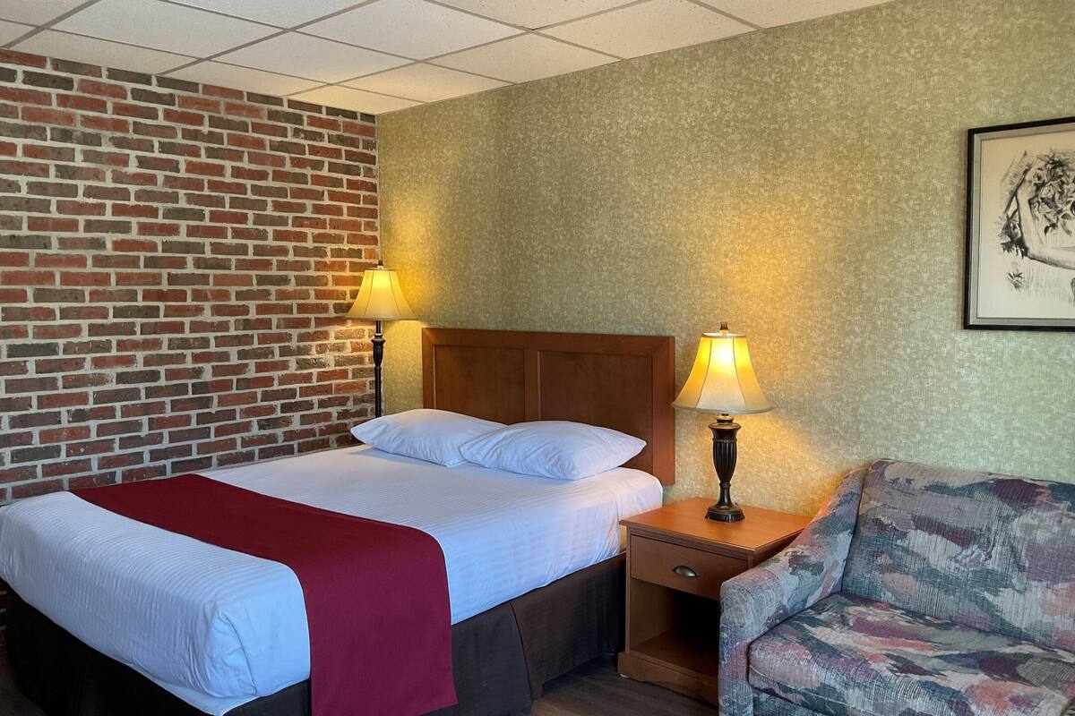 Hotellihuone, jossa on sänky, kaksi yöpöytälamppua, yöpöytä, tuoli, tiiliseinä ja seinällä oleva kehystetty kuva.