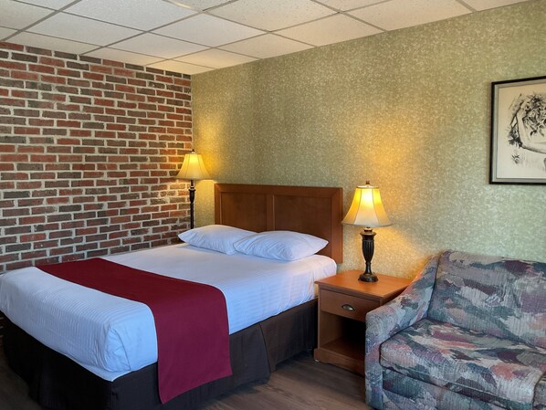 A hotel room with a bed, two bedside lamps, a nightstand, a chair, a brick wall, and a framed picture on the wall.