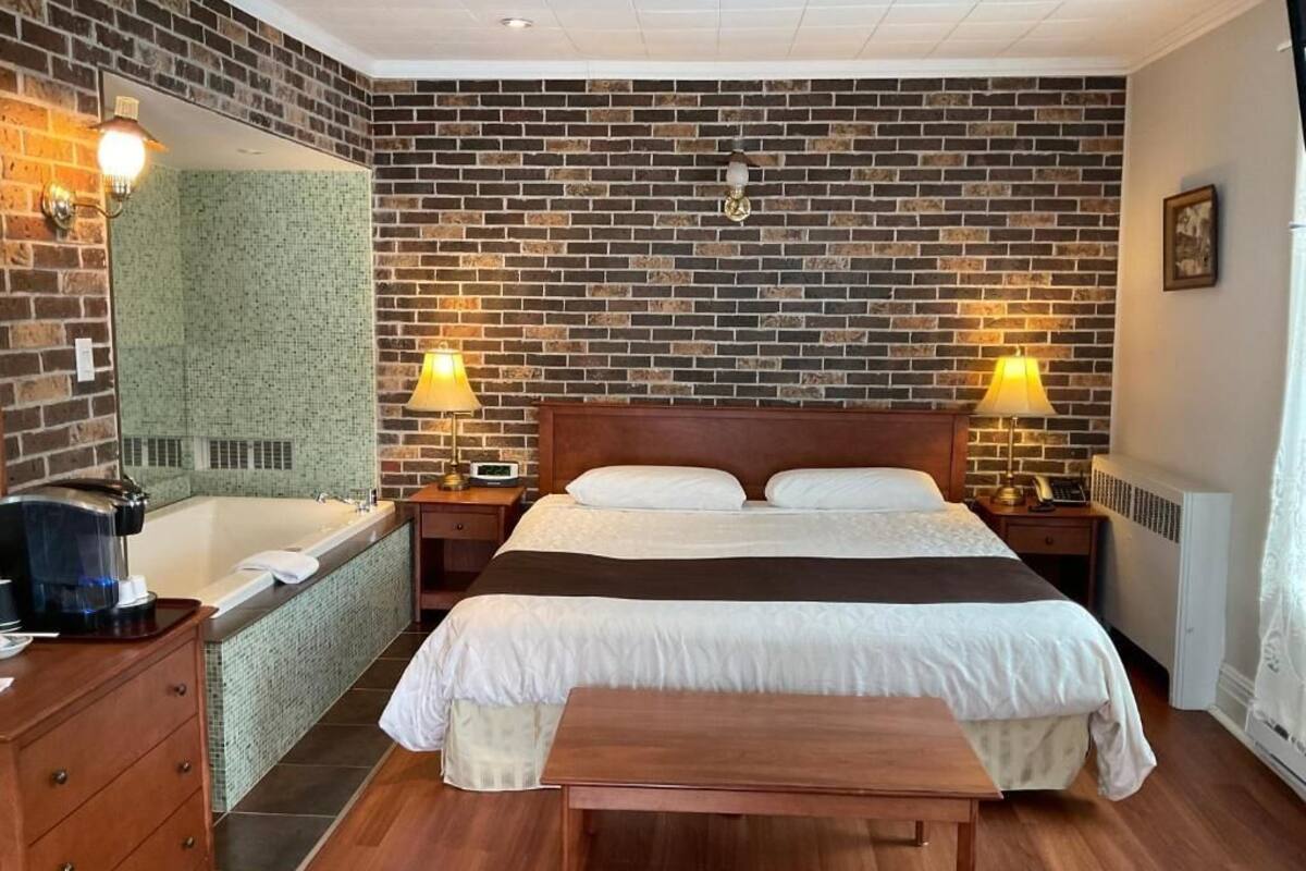 Hotellihuone, jossa on tiiliseinä, suuri sänky valkoisilla vuodevaatteilla, kaksi yöpöytälamppua, puinen penkki, kylpyammeella varustettu kylpyhuone ja peili.