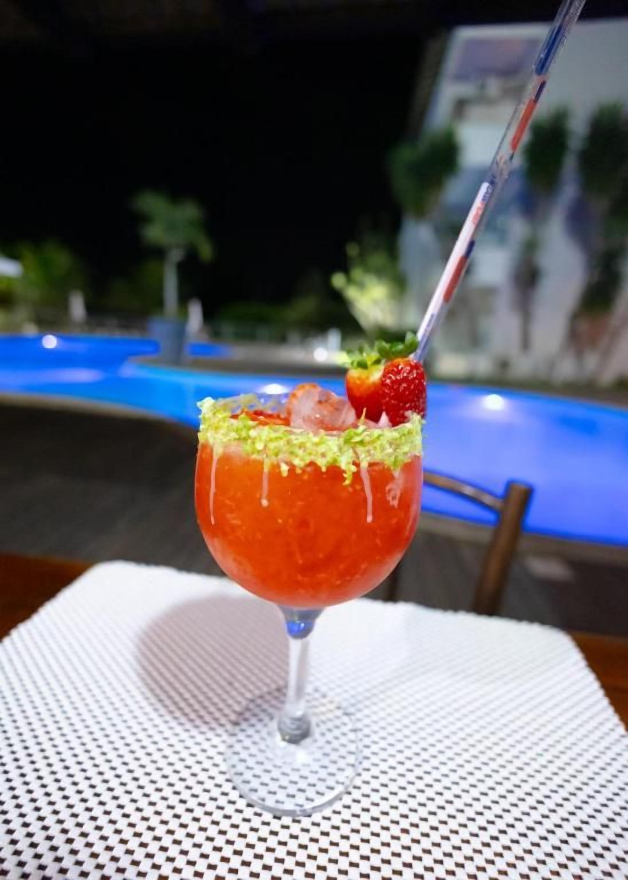 A cocktail in a glass with a straw, garnished with a strawberry and a green topping, placed on a table with a white napkin.