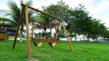 Un columpio de madera en un césped con árboles y un edificio al fondo.
