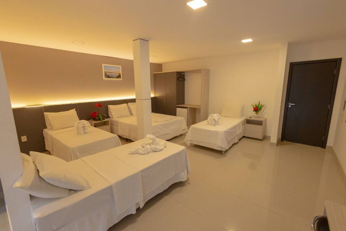 Une chambre d’hôtel avec trois lits, un bureau, une chaise, une petite table, une plante en pot et une porte.