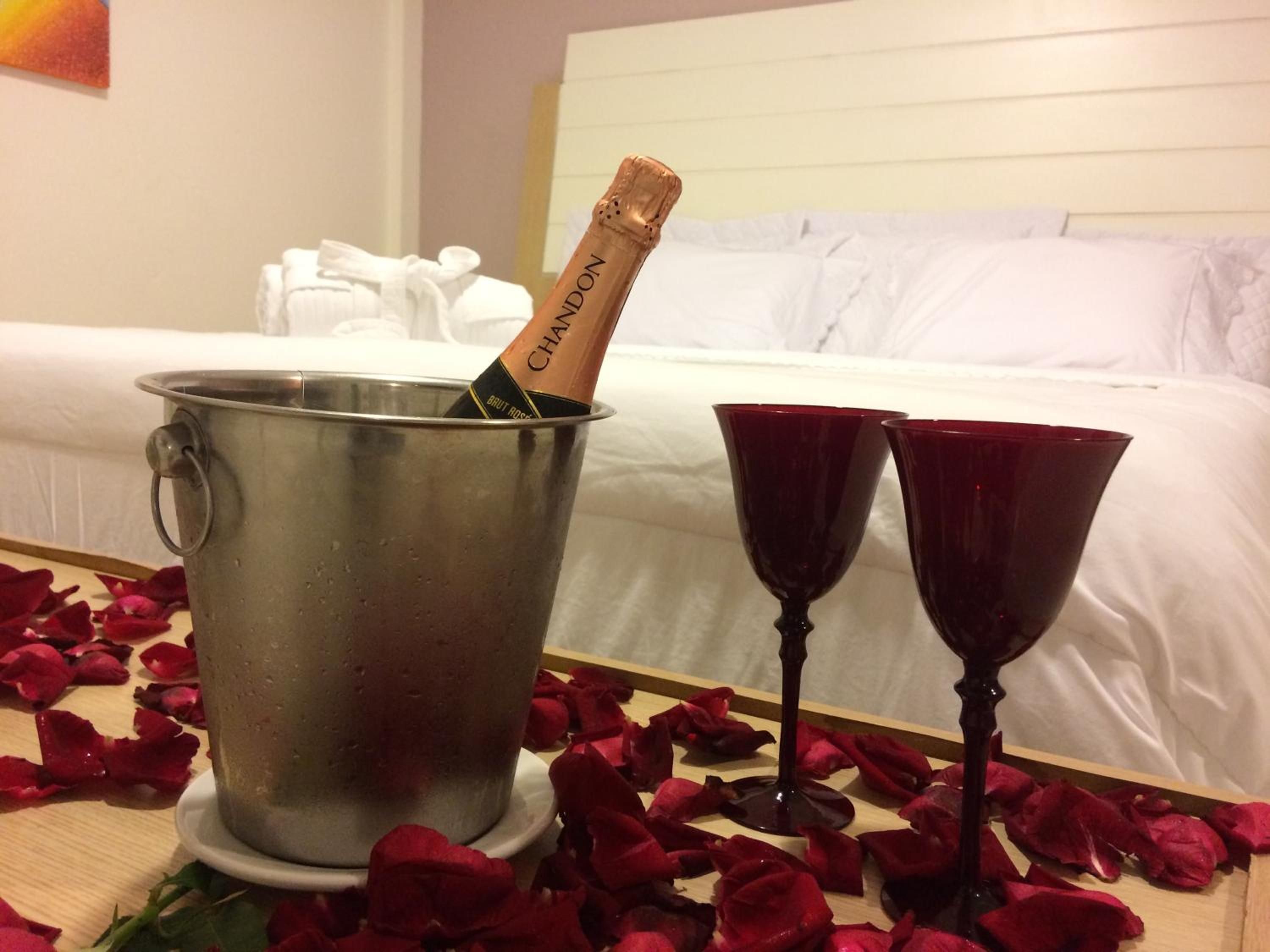 A bed with white linens, a metal bucket with a champagne bottle, and two red wine glasses on a table with rose petals.