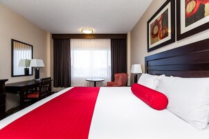 Quarto de hotel com uma cama grande, uma escrivaninha, uma cadeira, uma mesinha e uma janela com cortinas.