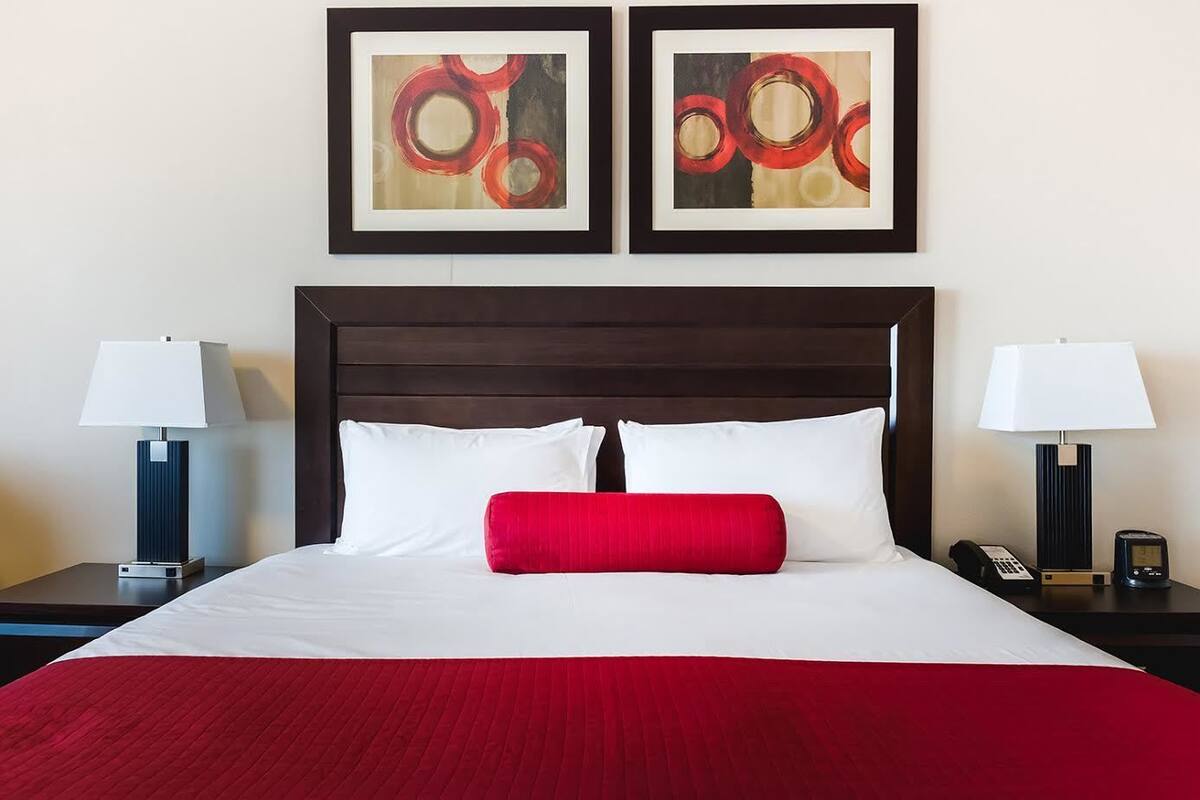 A hotel room with a large bed, white and red bedding, two framed abstract paintings above the bed, and bedside tables with lamps and phones.
