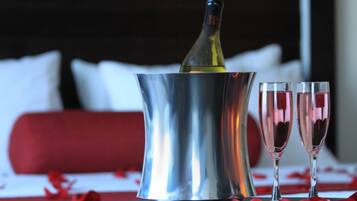 Una bottiglia di champagne in un secchiello metallico, due flute da champagne e petali di rosa rossa su un tavolo con un divano rosso e cuscini bianchi sullo sfondo.