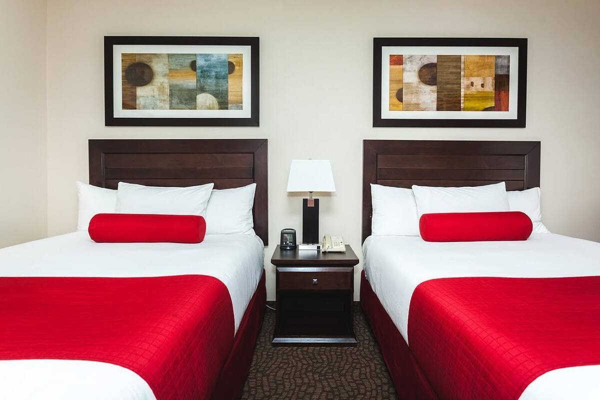A hotel room with two beds, each with white bedding and red accents, a nightstand with a lamp, and two framed abstract paintings on the wall.