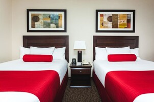Quarto de hotel com duas camas, cada uma com roupa de cama branca e detalhes vermelhos, uma mesa de cabeceira com abajur, e duas pinturas abstratas emolduradas na parede.