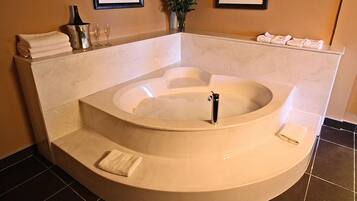 A large, round bathtub with a built-in seat, a small sink, and a bottle of champagne. There are towels stacked on the side and a vase with red flowers.