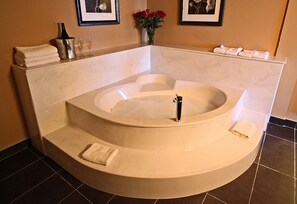 Una bañera grande y redonda con asiento integrado, un lavamanos pequeño y una botella de champán. Hay toallas apiladas a un lado y un jarrón con flores rojas.