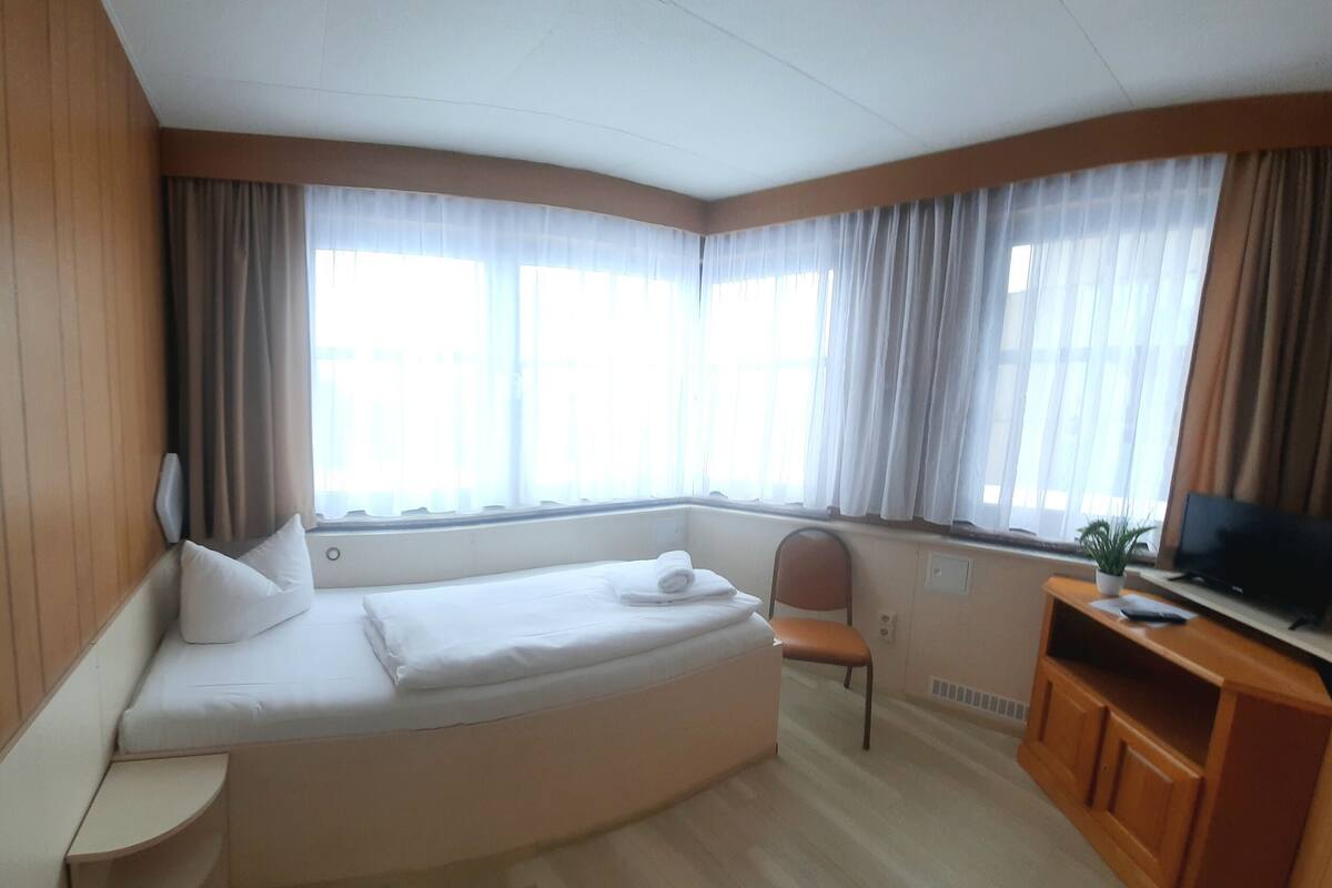 Ein Hotelzimmer mit Bett, Sessel, Fernseher und einem Fenster mit Vorhängen.