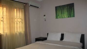 A bedroom with a bed, a nightstand, an air conditioning unit, and a window with curtains.