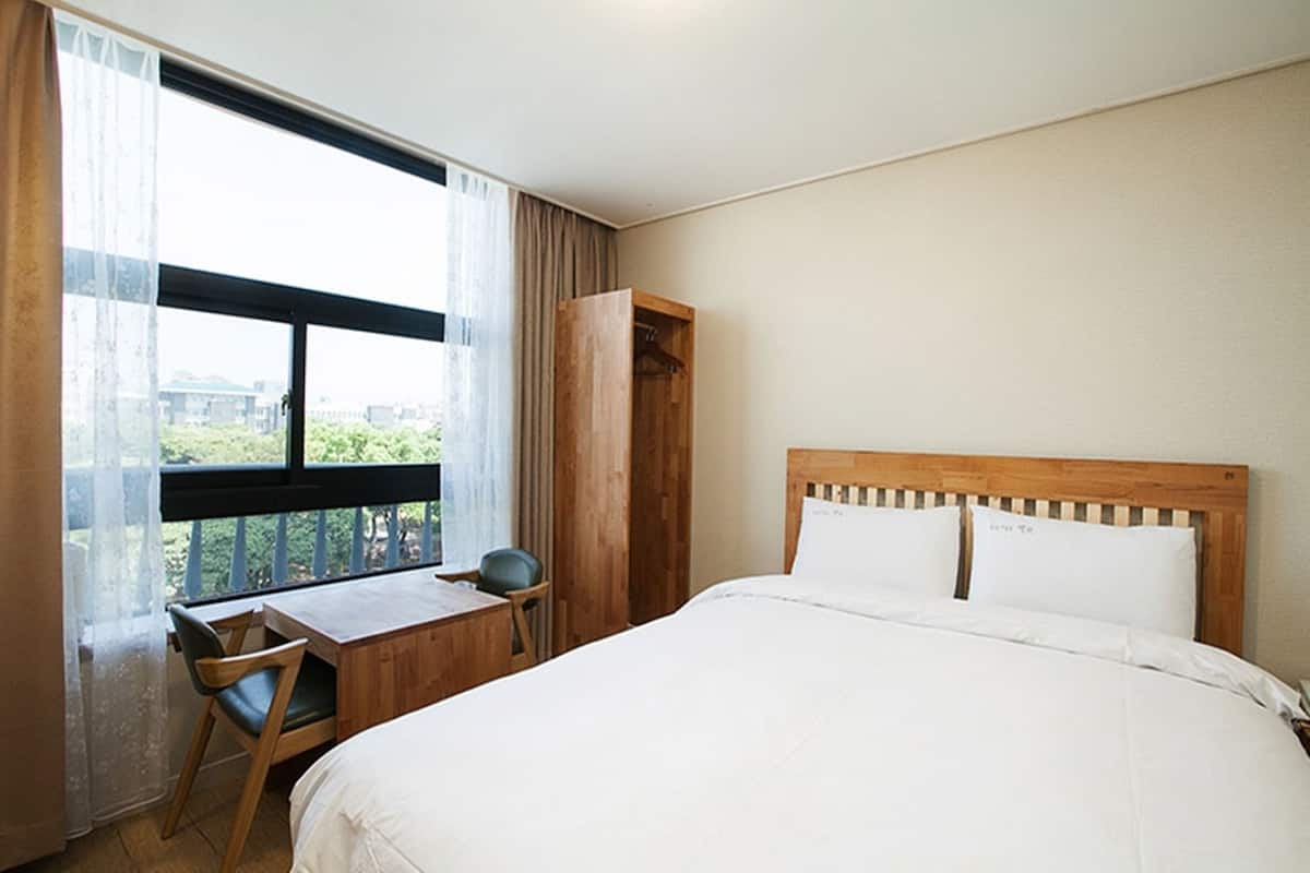 Une chambre d’hôtel avec un lit, un bureau et une chaise. Il y a une fenêtre avec des rideaux et une vue sur les bâtiments à l’extérieur.