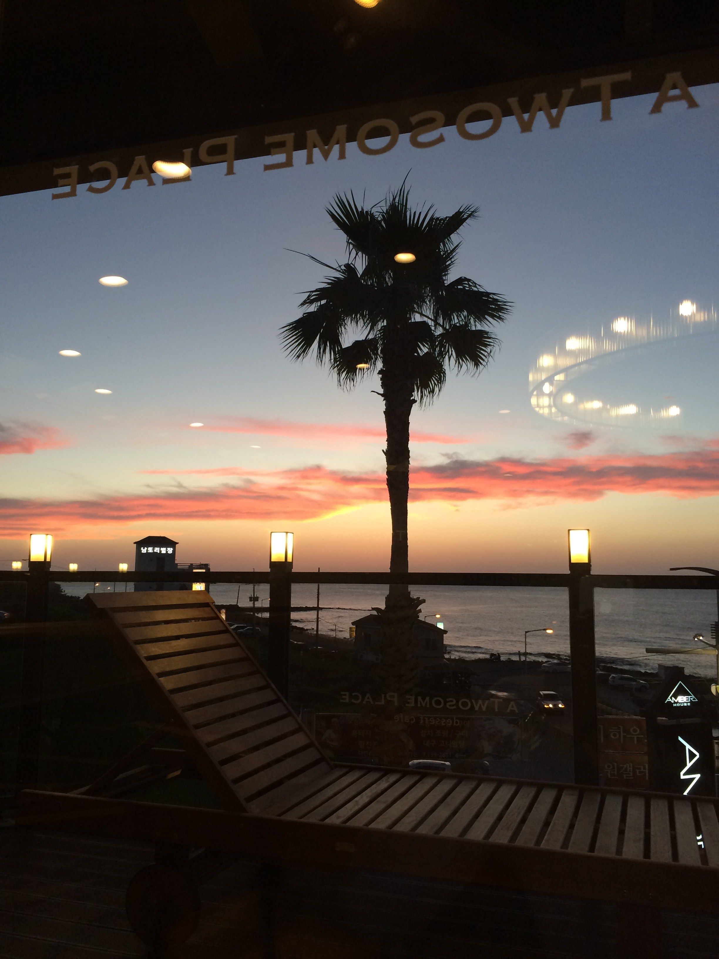 Une vue sur le coucher de soleil depuis un balcon agrémenté d’un palmier, d’une terrasse en bois et d’un pont situé au loin.