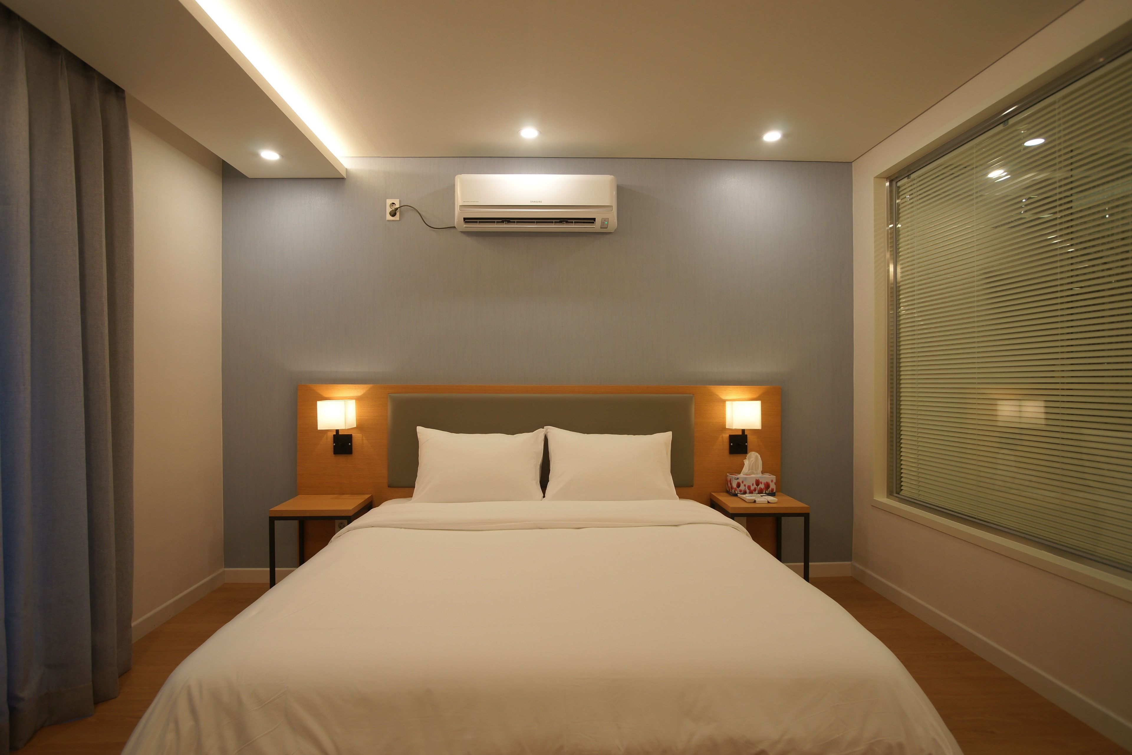 Habitación de hotel con una cama grande, dos lámparas de noche, un equipo de aire acondicionado y una ventana con persianas.