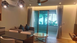 A modern living room with a sofa, a dining table, and a TV. There is a balcony with curtains and a view of greenery. - The Regent Residence Kamala (Kamala)