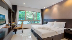 A modern hotel room with a large bed, a desk with a computer, and a window with a view of trees. - The Regent Residence Kamala (Kamala)