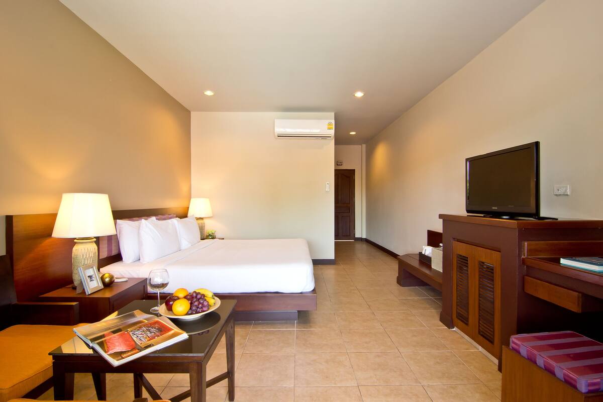 A hotel room with a bed, bedside lamps, a television, a table with a magazine and fruit, and an air conditioning unit.