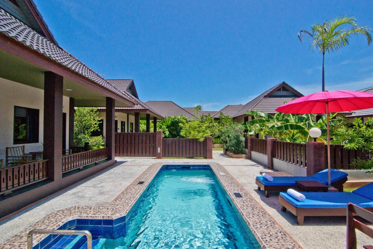 A swimming pool with lounge chairs and umbrellas, surrounded by buildings with tiled roofs and a wooden fence.