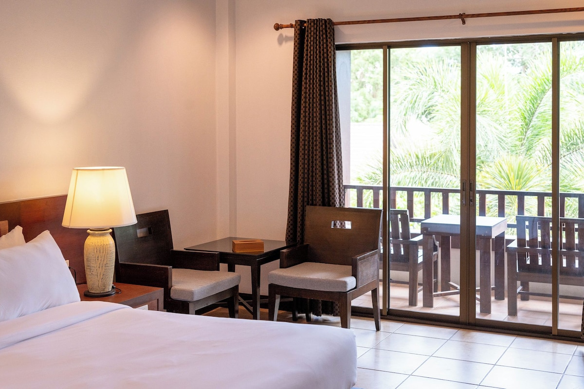 A hotel room with a bed, a desk, a chair, a table, and a balcony with outdoor furniture.