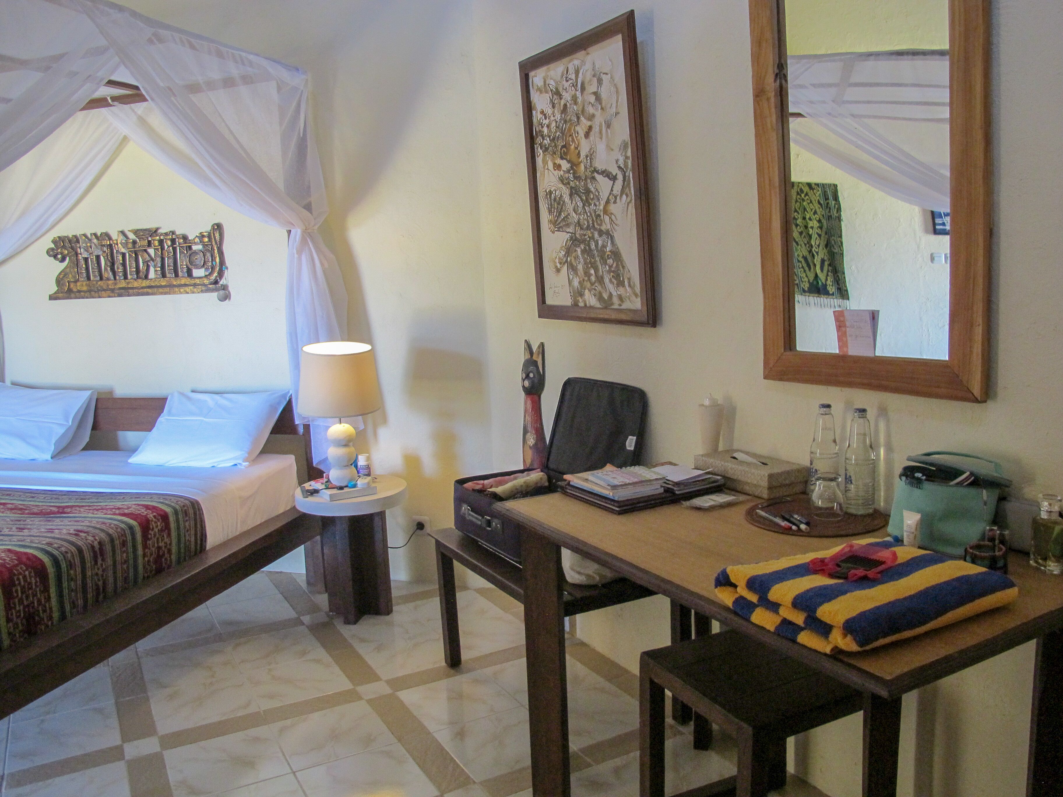 A hotel room with a bed, a desk, a chair, a lamp, a mirror, and a painting on the wall.