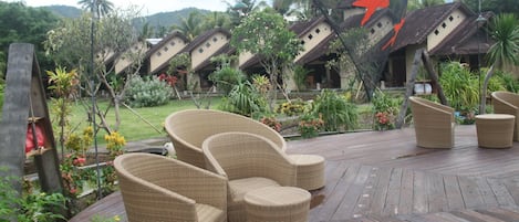 Eine Holzterrasse mit Korbmöbeln, ein tropischer Garten und traditionelle Häuser mit Strohdächern.