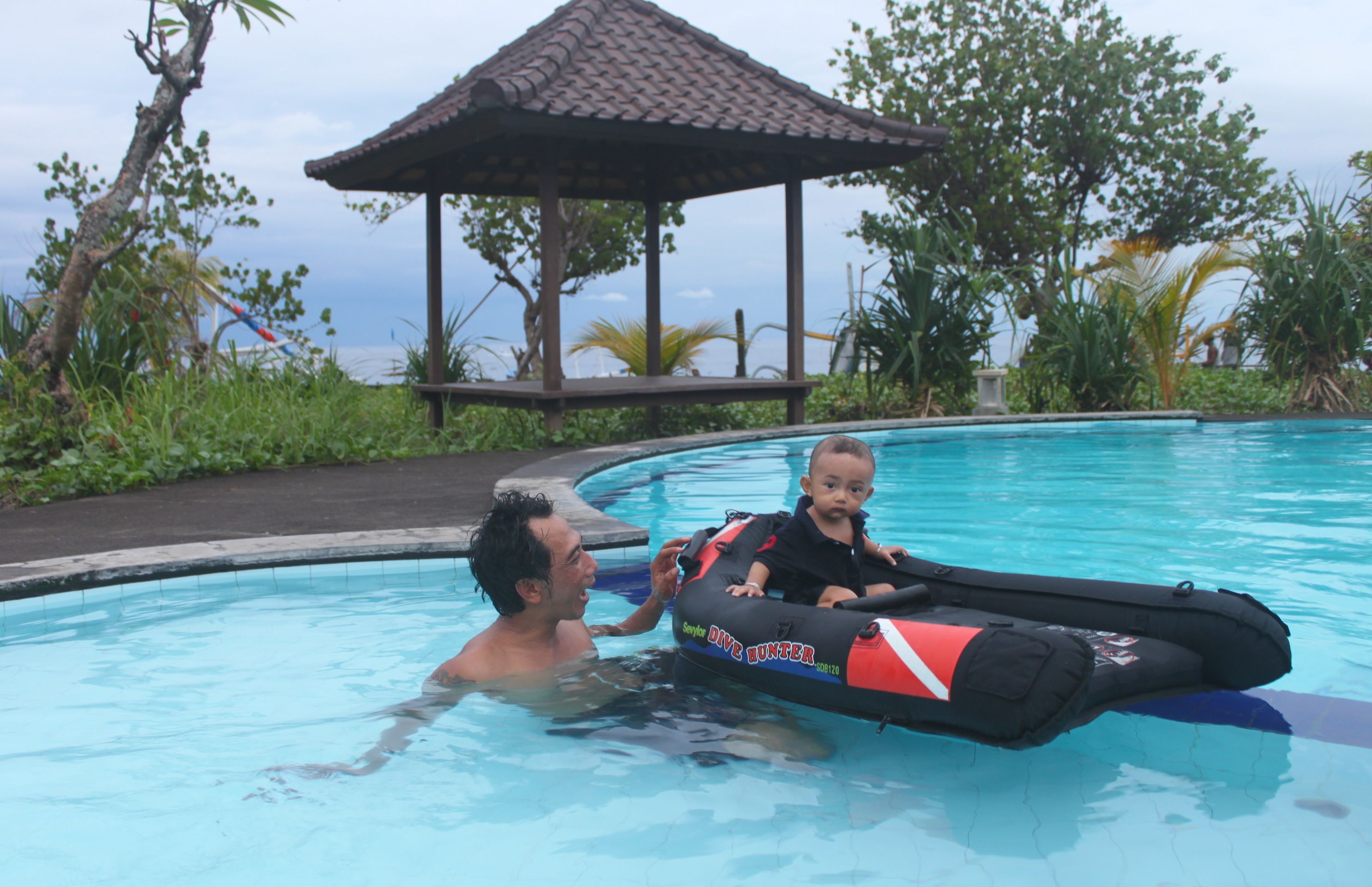 A man and a child in a swimming pool with a floating device, surrounded by greenery and a gazebo.