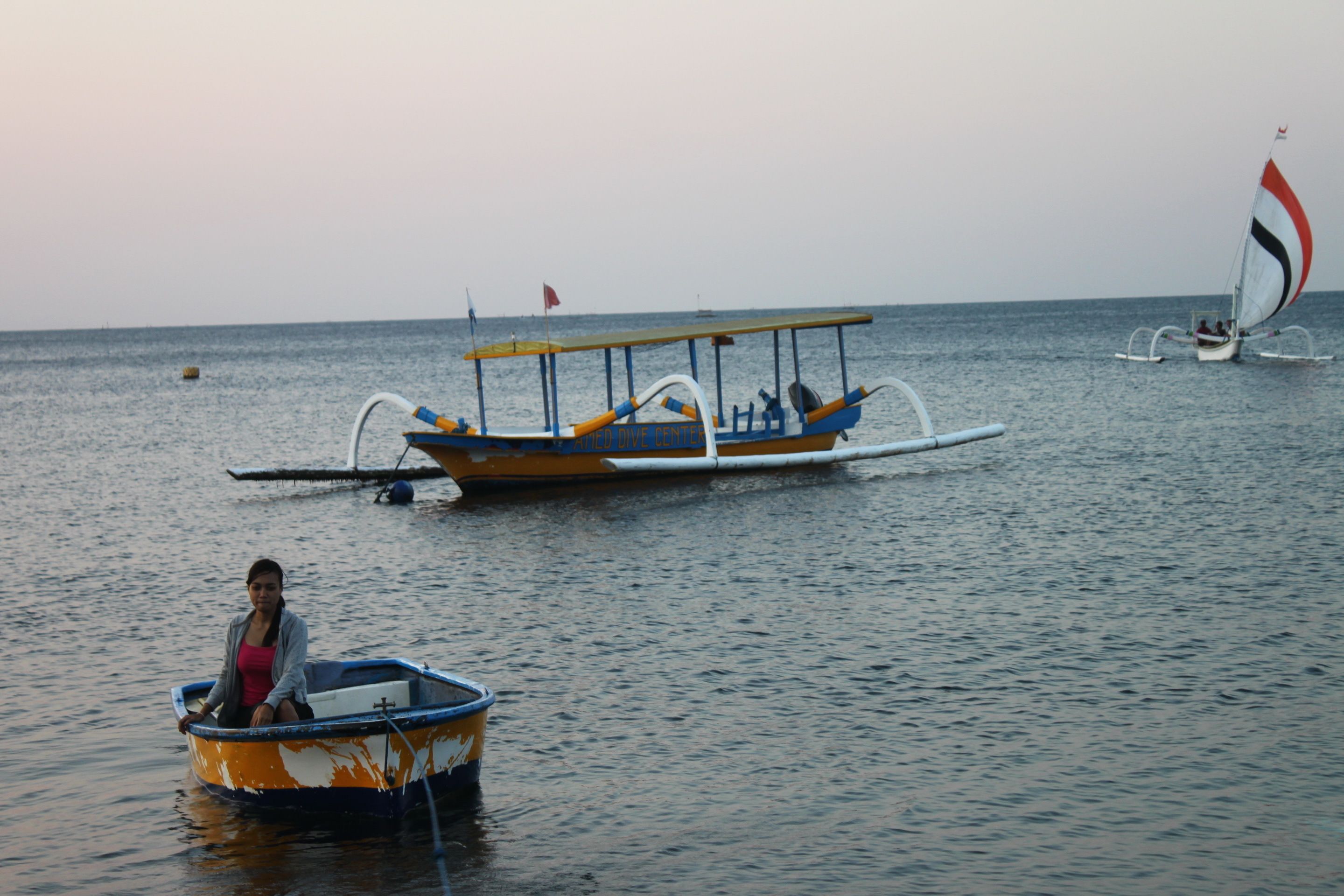 A small boat with a person on it, a larger boat with a sail in the background, and calm water.
