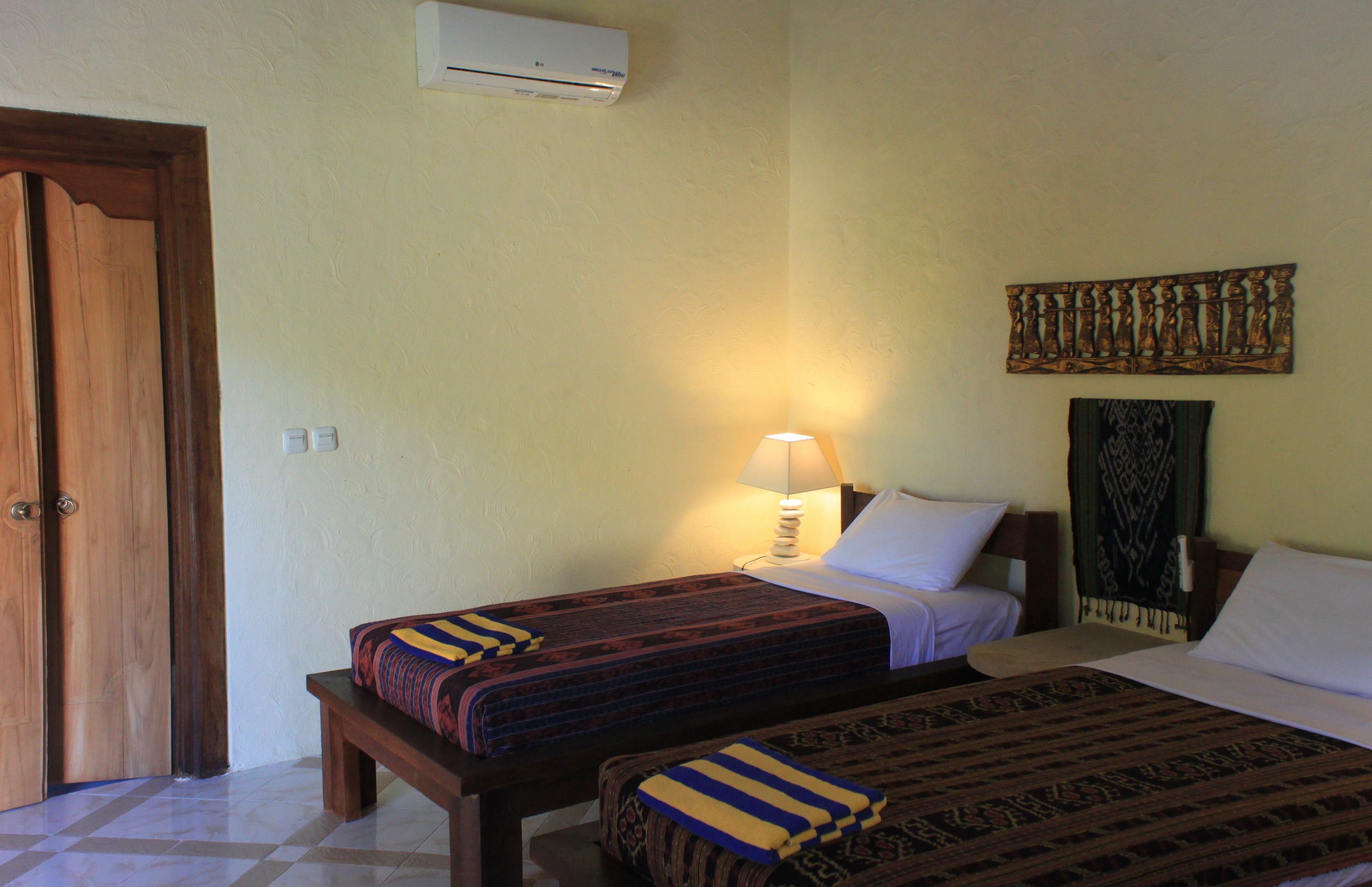 A hotel room with two beds, a nightstand with a lamp, a wall-mounted air conditioner, and a door.