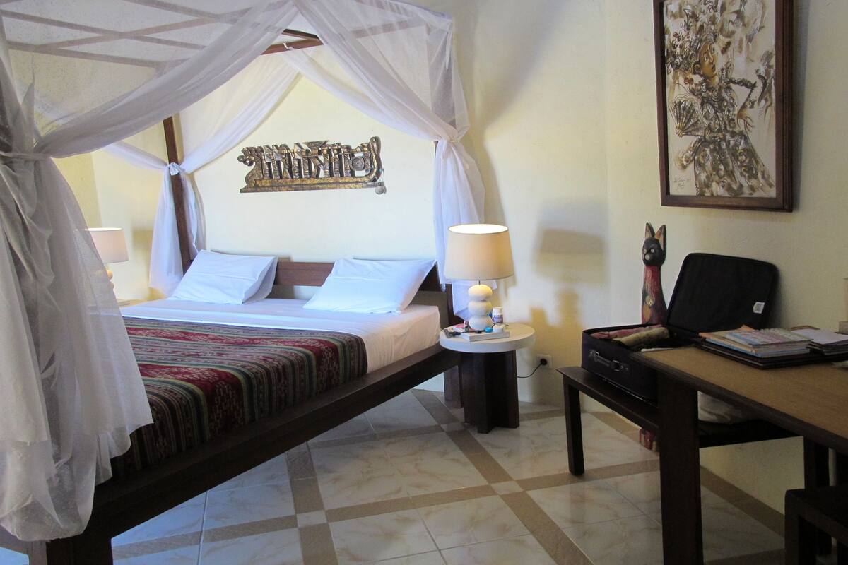 A room with a bed under a canopy, a desk with a laptop, a chair, and a wall with a decorative piece.