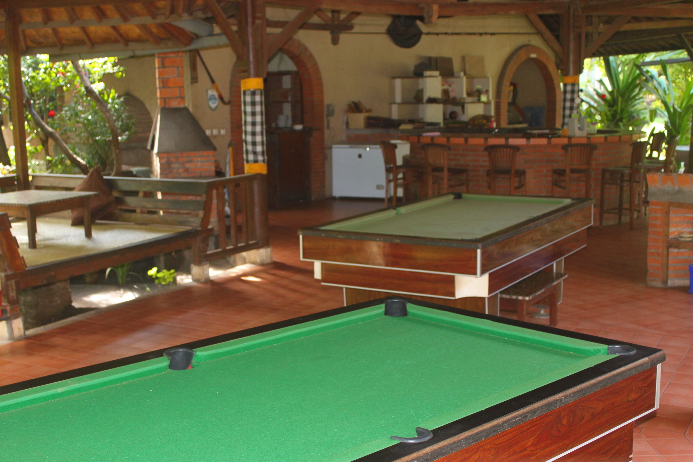 A pool table in a covered outdoor area with a brick fireplace, wooden furniture, and a kitchen area in the background.