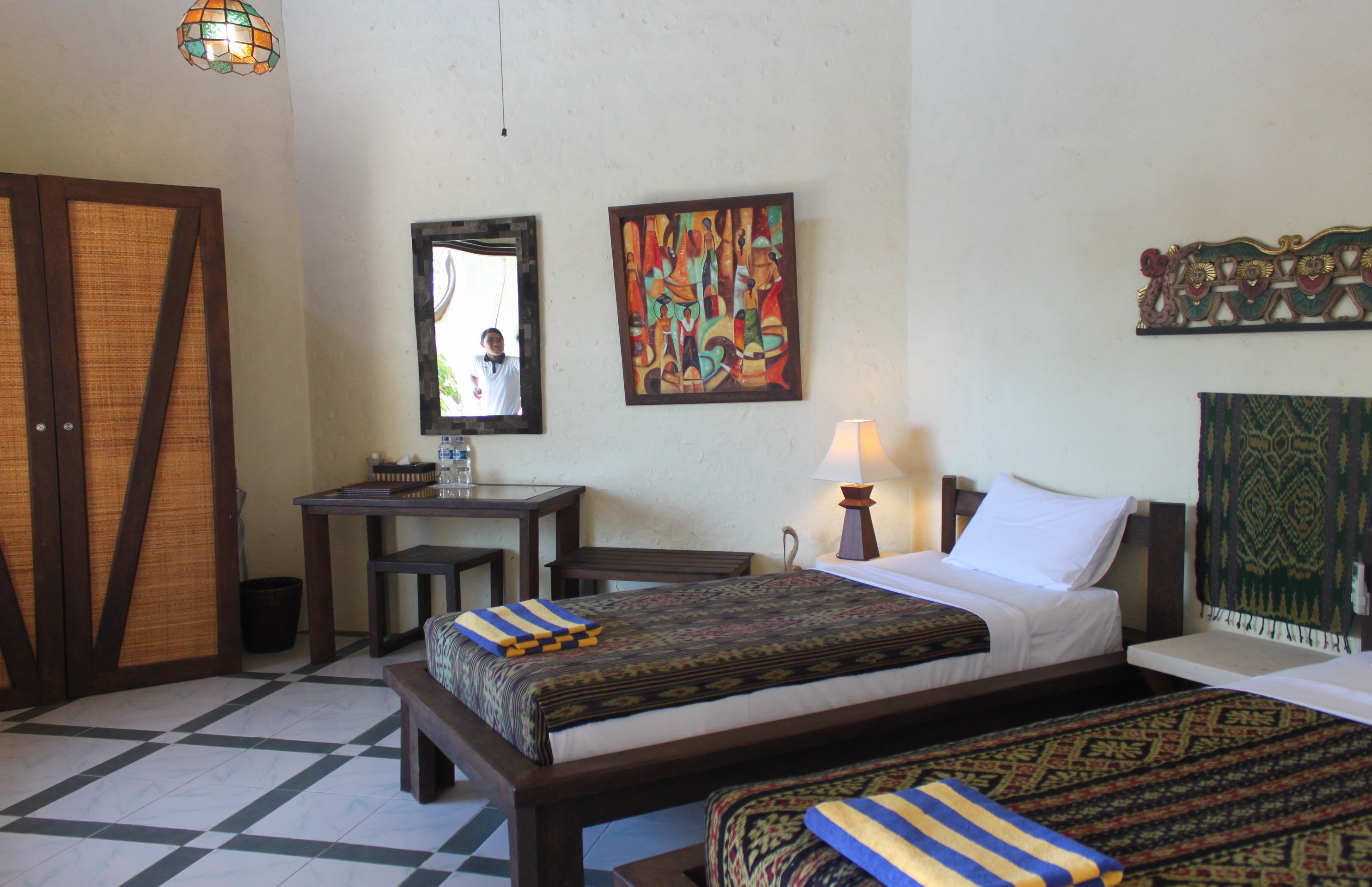 Une chambre d’hôtel avec deux lits, un bureau, une chaise, une lampe et des œuvres d’art accrochées aux murs.