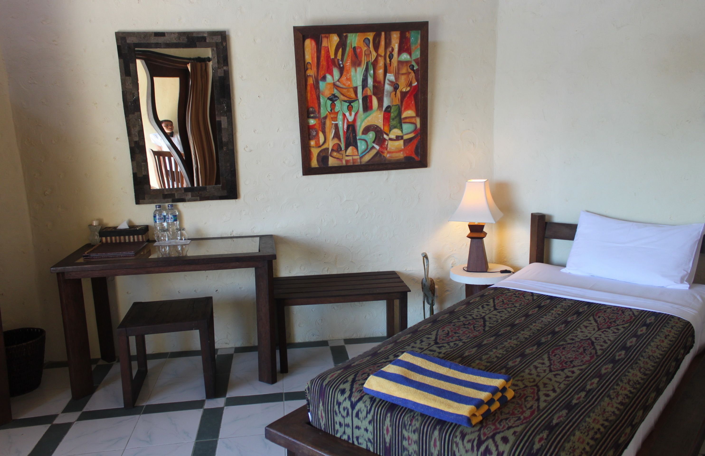 A hotel room with a bed, a desk, a chair, a lamp, and a painting on the wall.