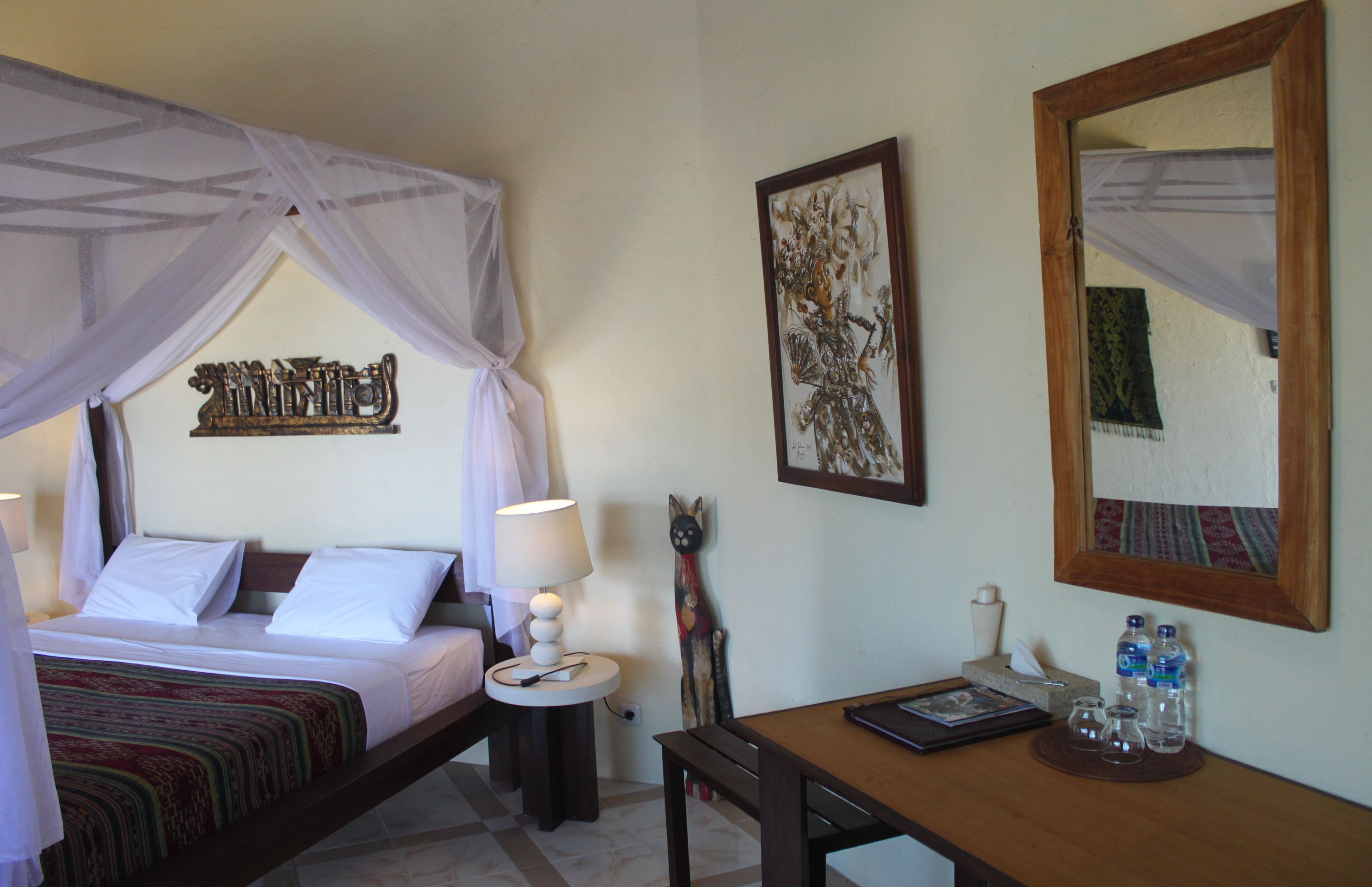 A hotel room with a bed under a canopy, a bedside table with a lamp, a small desk with a chair, and a wall-mounted mirror.