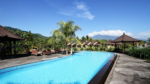 A swimming pool with lounge chairs and thatched-roof structures, surrounded by tropical vegetation and mountains. - Hotel Uyah Amed & Spa Resort (Karangasem)