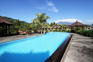 A swimming pool with lounge chairs and thatched-roof structures, surrounded by tropical vegetation and mountains.