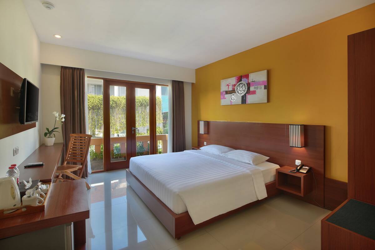 A hotel room with a large bed, a desk with a chair, a TV, and a balcony with curtains.