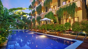 A swimming pool with lounge chairs and umbrellas, surrounded by lush greenery and a multi-story building with balconies. - Bali Chaya Hotel Legian (Legian)