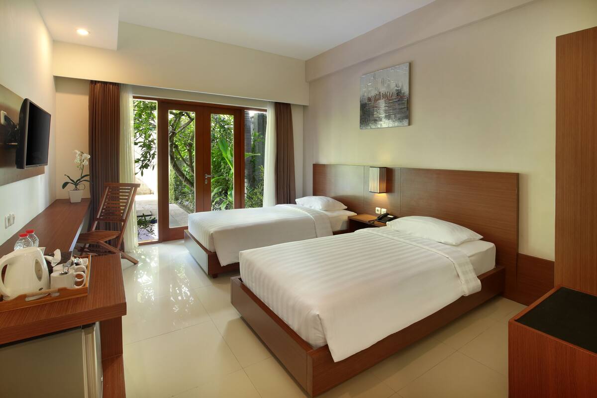 A hotel room with two beds, a desk with a TV, a coffee maker, and a view of greenery outside.