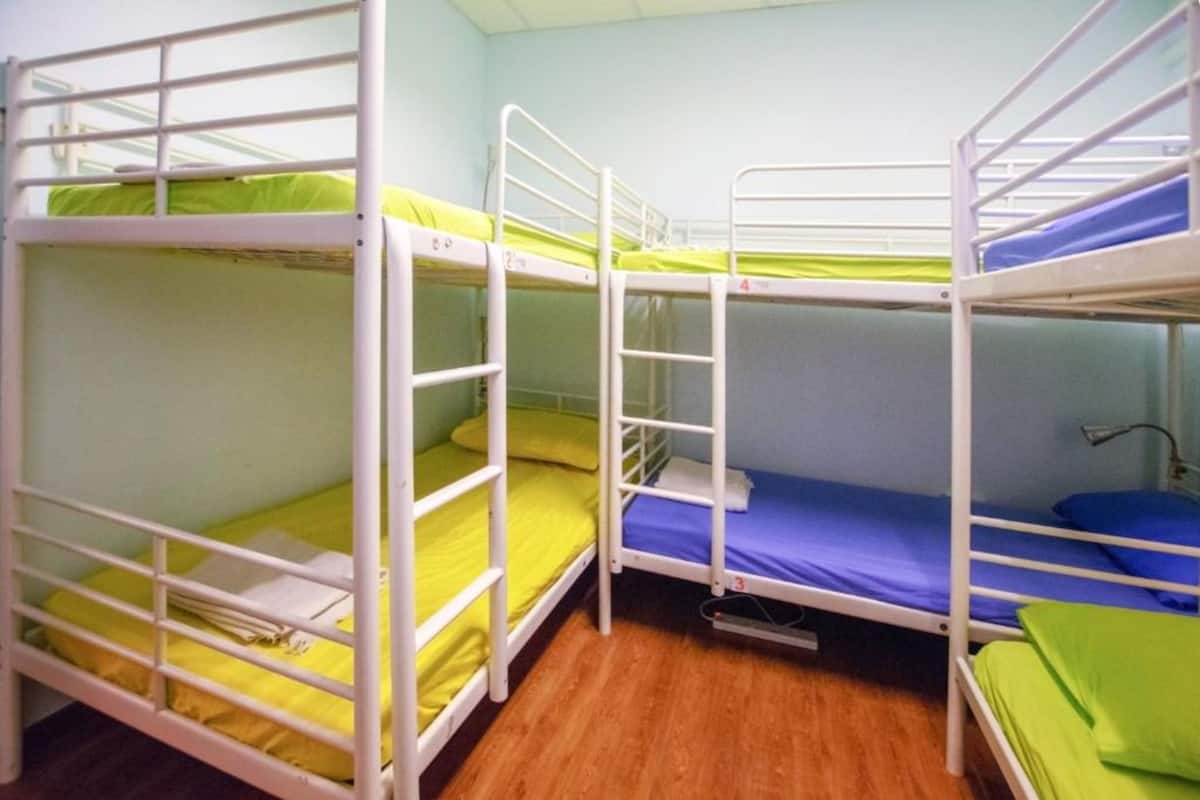 A dormitory room with bunk beds, wooden flooring, and a single bedside table with a lamp.