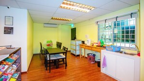 A bright, well-lit room with a wooden table and chairs, a window with blinds, a small kitchenette, and a shelf with magazines. - Vintage Inn @ Clarke Quay - Hostel (Singapore)