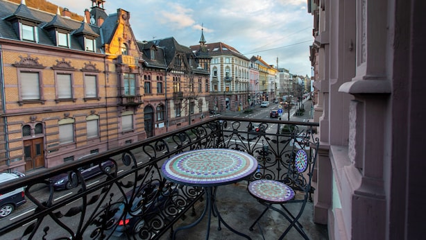 Un balcón con barandilla de hierro forjado y dos mesas con cubierta de mosaico, con vistas a una calle con edificios históricos y cielo despejado.
