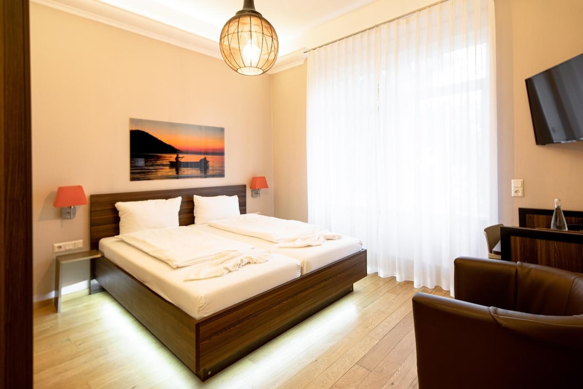 Quarto de hotel com uma cama grande, uma pintura de um pôr do sol, uma televisão na parede e uma cadeira.