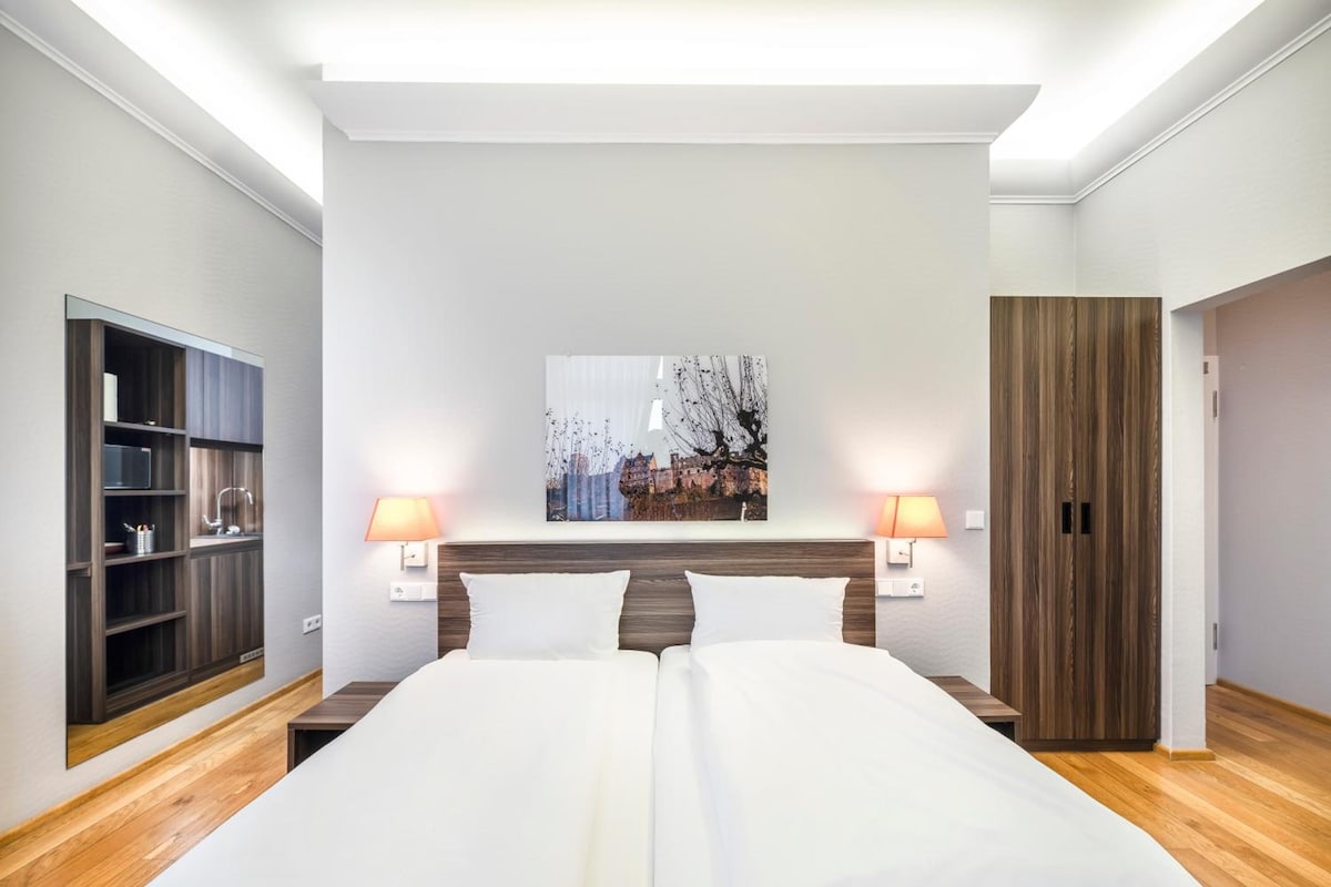 Quarto de hotel com cama, duas luminárias de cabeceira, um guarda-roupa e uma obra de arte na parede.