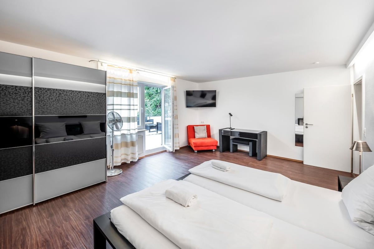 Quarto moderno com cama, uma cadeira vermelha, TV e porta de correr para uma varanda.