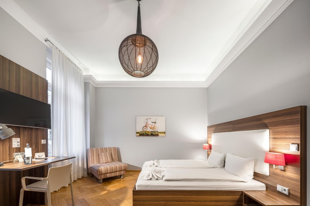 Quarto de hotel moderno com piso de madeira, cama com roupa branca, escrivaninha com cadeira, poltrona listrada, TV na parede e luminária pendente.