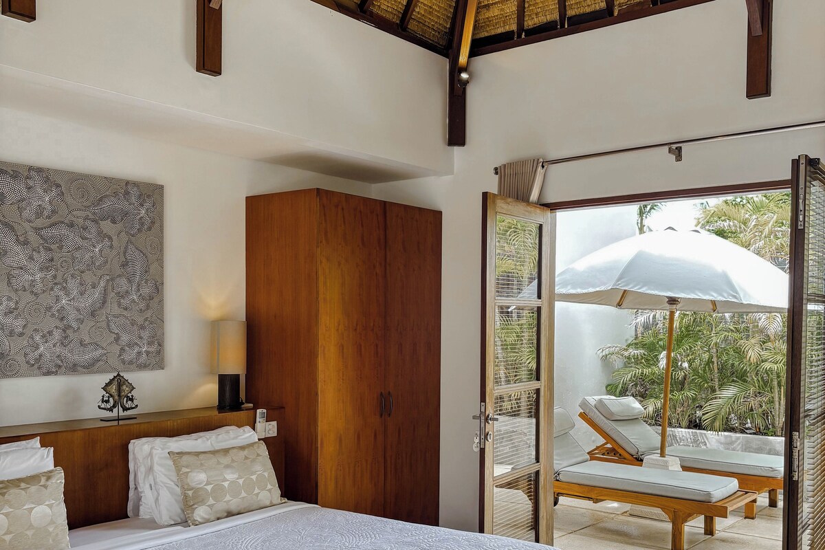 Quarto com cama, guarda-roupa de madeira e uma janela grande com vista para espreguiçadeiras ao ar livre.
