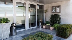 Entrada de hotel con puertas de vidrio, un bote de basura, plantas en macetas y una pequeña mesa con lámpara.