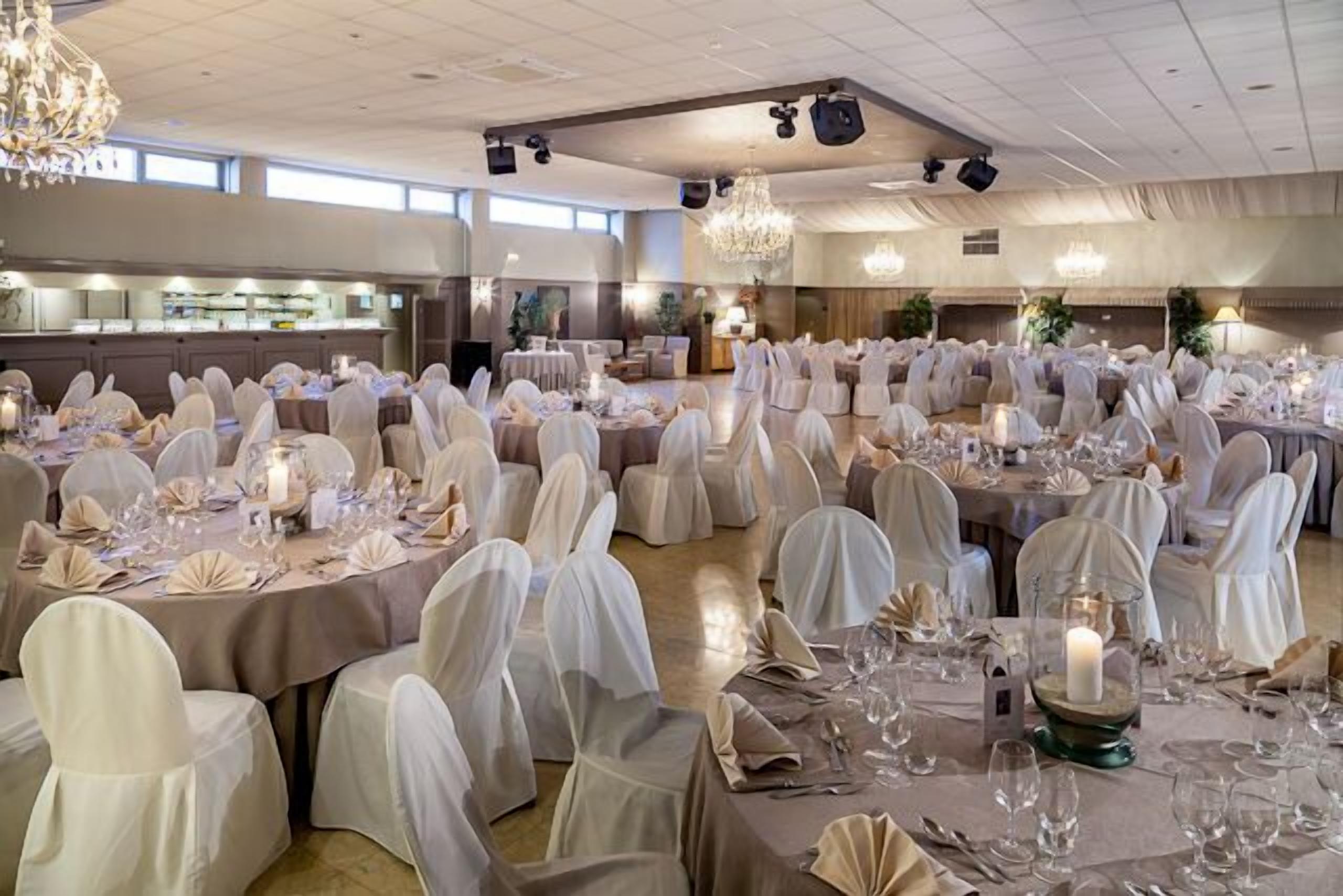 Una sala da banchetto con tavoli rotondi apparecchiati per un evento formale, con lampadari e una cucina sullo sfondo.