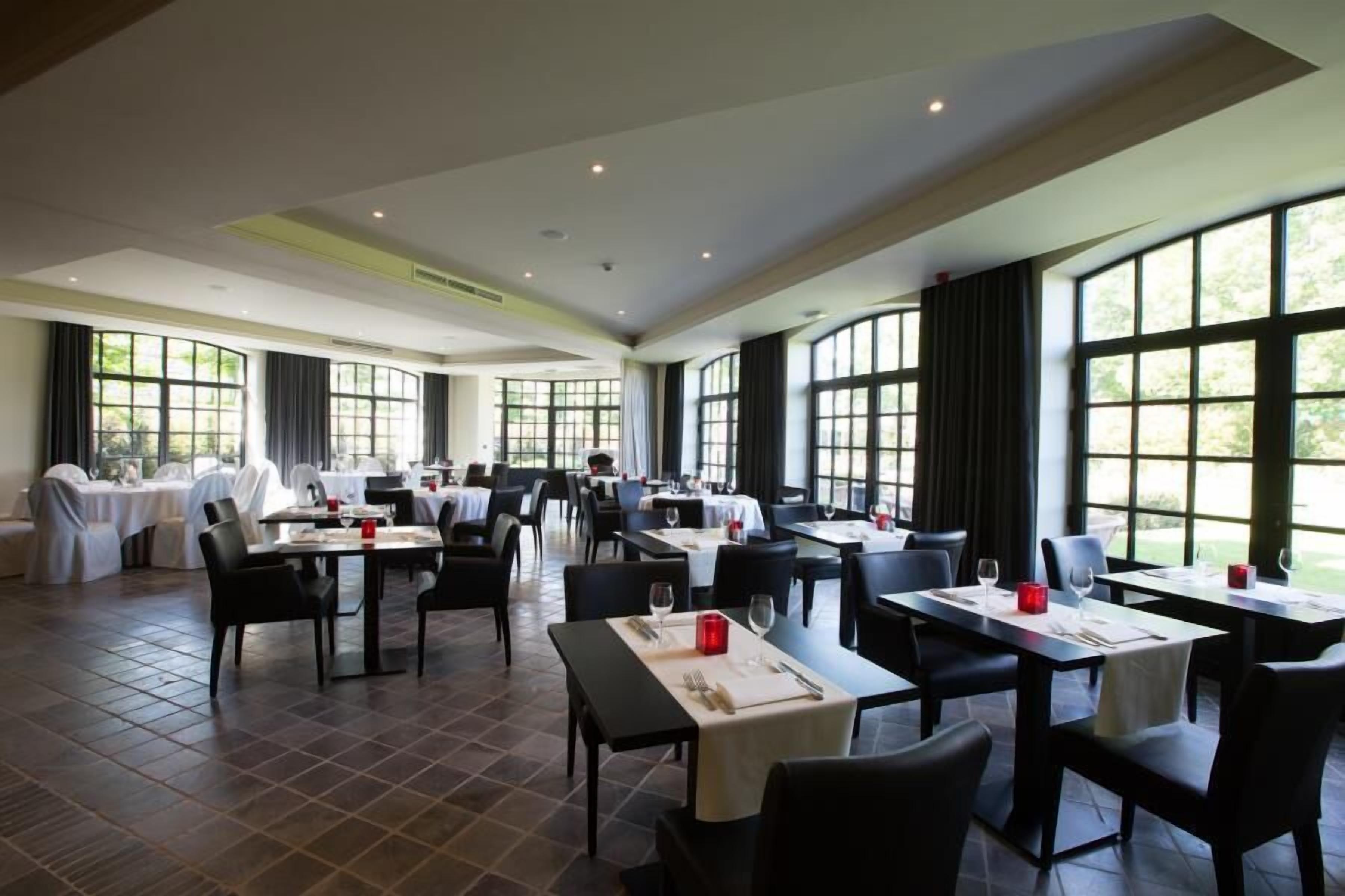 A spacious dining area with multiple tables and chairs, large windows offering views of the outdoors, and a high ceiling with recessed lighting.